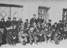 Duża grupa mężczyzn w czapkach-leninówkach siedzi na tle budynku.