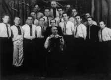 Grupa mężczyzn w koszulach-rubachach, jeden z harmonią (zespół, chór?).