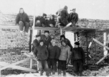 Mężczyźni przy budowie domu. Grupa mężczyzn w strojach roboczych stoi i siedzi na rusztowaniach przy ceglanym murze.