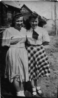 Dwie młode kobiety w letnich strojach, stojące na tle drewnianych zabudowań.