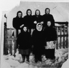 Siedem kobiet (dziewczynek?) w zimowych strojach i chustkach, stoją przy drewnianym płocie.