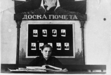 Dziewczyna siedząca za stołem nad książką. Za nią tablica z fotografiami, profilem Lenina i napisem: Doska pocziota.