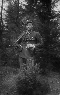 Portret mężczyzny w mundurze z bronią na tle lasu.