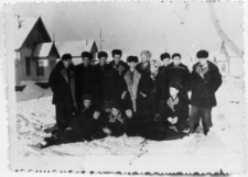 Grupa os&oacute;b w zimowych ubraniach na tle drewnianych domk&oacute;w z gankami. Polacy, więźniowie obozu D-2. Od lewej stoją: Hipolit Suchocki, Wacław Osipowicz, NN, Alfons Nadziejko, Bolesław Peckiel, Antoni Rymsza, NN, NN, Jerzy R&oacute;żanowski, NN; pierwszy od prawej siedzi Antoni Witukiewicz.