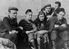 Grupa osób w strojach letnich, siedzą na tle krajobrazu.