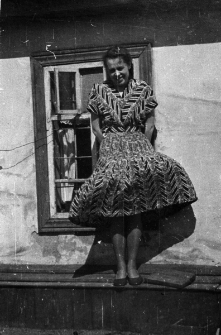 Janina Durlik - w sukience, stoi na tle budynku, przy oknie.