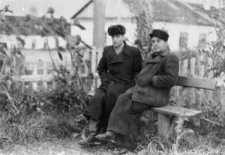 Dw&oacute;ch mężczyzn na ławeczce na tle budynk&oacute;w i krzew&oacute;w.