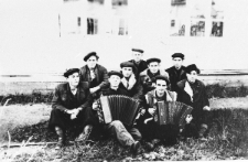 Grupa mężczyzn, dw&oacute;ch z harmoniami, na tle budynku.