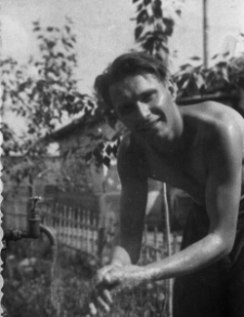 Mężczyzna rozebrany do pasa, myjący ręce. W tle zarośla i niski budynek.