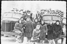 Grupa ludzi w waciakach między dwiema ciężarówkami załadowanymi drewnem.