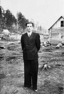 Mężczyzna w garniturze, w tle las i drewniany budynek. Tadeusz Bukowy na zesłaniu. Zdjęcie wykonane w latach 50-tych.