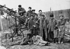 Wolnoosiedleńcy oraz Ukraińcy deportowani w 1950 r. Grupa ludzi w strojach roboczych przy metalowej maszynie (szkielet taśmociągu?). W tle ogrodzenie.