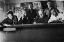 Klasa szkolna. Czterech mężczyzn w ławce, dw&oacute;ch stoi za nimi. W tle plansze na ścianie.