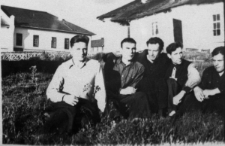 Grupa mężczyzn siedzących na trawie przed budynkami. Pierwszy z lewej: Eryk Barcz (Lech Kożuchowski).