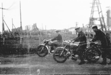 Mężczyźni na motocyklach, w tle baraki dawnego obozu.