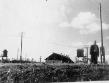 Karol Prass, zwolniony z łagru, na tle opuszczonego obozu (barak, ogrodzenie, wieżyczki strażnicze).