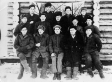 Grupa mężczyzn w waciakach przed budynkiem z drewnianych bali, za nimi wisi namalowany leśny widoczek, prawdopodobnie są to więźniowie [byli?] łagrów w ZSRR, 1955-1956.
