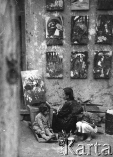 Aniela Wzorek malująca obrazki (ilustracje do bajek dla przedszkoli), na ścianie wiszą gotowe prace, obok bawią się dzieci Zygmunt i Danuta; lata 1944-46.