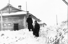 Dwaj mężczyźni w zimowych ubraniach stoją na śniegu przed domem, prawdopodobnie są to Polacy zesłani do ZSRR.