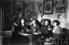 Grupa osób w saloniku, z lewej na oparciu fotela siedzi Janusz Owsianny, obok niego w fotelu siedzi jego matka Apolonia, dalej młody mężczyzna NN, doktorowa Kleinowa (siostra Apolonii) i młoda kobieta NN.